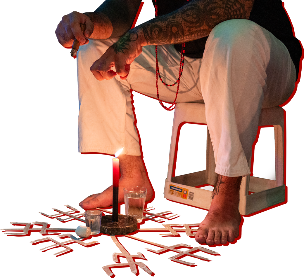 Pessoa incorporada com Exu sentada em um banquinho, vestindo calça branca e usando colar de contas. Os pés estão descalços próximos a uma vela vermelha acesa sobre uma base preta, cercada por tridentes brancos desenhados no chão. O braço exposto exibe tatuagens, enquanto a mão segura um objeto semelhante a um charuto. O fundo é vermelho, destacando a cena ritualística.